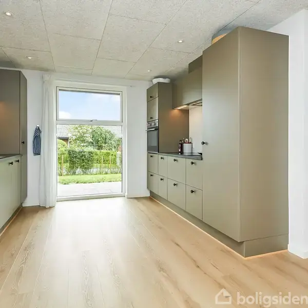 Et moderne køkken med grønne skabe langs begge vægge. Sollys skinner gennem en glasdør, der fører til en have. Trægulv og et farverigt kunstværk på væggen.