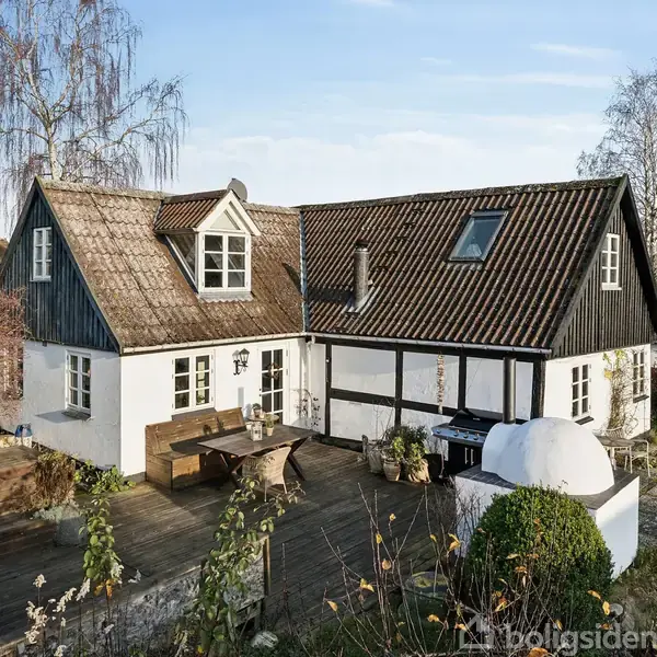 Et charmerende bindingsværkshus med sort træværk og hvidkalkede vægge står stille i en have. En træterrasse med borde og bænke omgiver huset, mens efterårstræer danner baggrund.