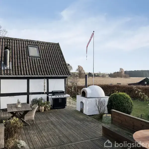 Terrasse med træmøbler og grill står foran et hvidt bindingsværkshus. En pizzaovn er ved siden af, flagstang med dannebrog i baggrunden, og marker omgiver området.