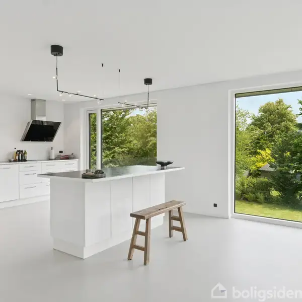 Et moderne, lyst køkken med et hvidt køkkenbord, hvorpå der står en dekorativ genstand. En bænk står ved siden af. Store vinduer giver udsigt til en frodig have med træer.