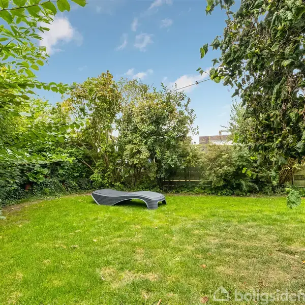 En sort liggestol står på en grøn græsplæne omgivet af tætte buske og træer, under en blå himmel med få skyer.