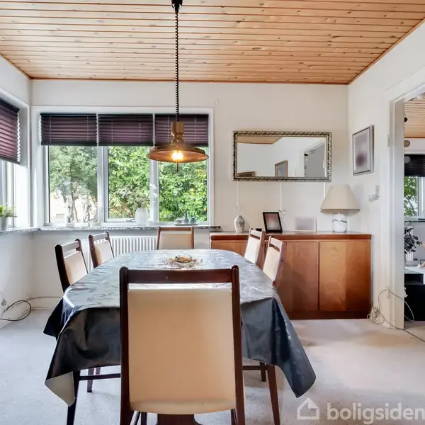 Spisebord med seks stole omgivet af hvidmalede vægge og træloft. En lampe hænger over bordet. Store vinduer til venstre med udsigt til grønne træer.