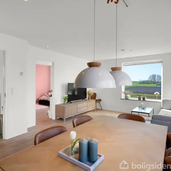 Spisebord med stearinlys står i forgrunden, omgivet af brune stole. I baggrunden ses en grå sofa ved vindue, med TV på et lavt skab. Døren fører til et lyserødt rum.
