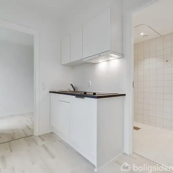 Et køkkenområde med hvide skabe og en sort bordplade. En åbning til en flisebelagt badeværelse med spejl og vask. Lyse trægulve i begge rum. Ingen tekst fundet.