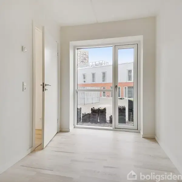 Et lyst rum med trægulv og en åben dør mod en lille balkon. Store vinduer viser udsigt til en bygning med fladt tag og udenfor er en mindre terrasse.