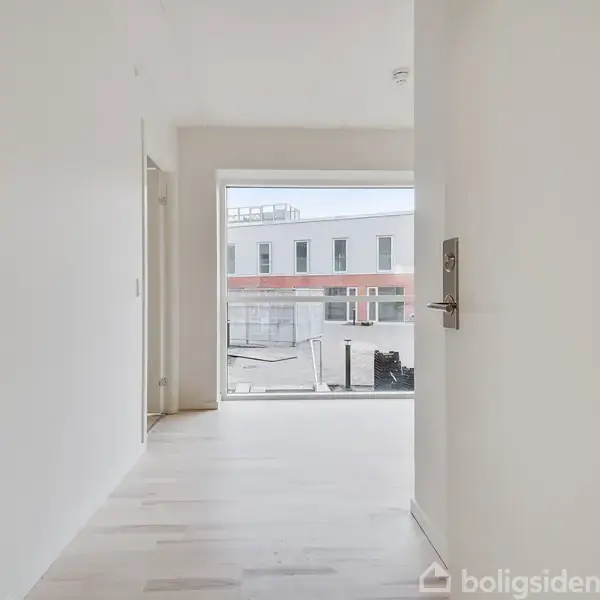 Lys dør i hvid korridor åbner mod altan med udsigt til moderne bygning på tags.