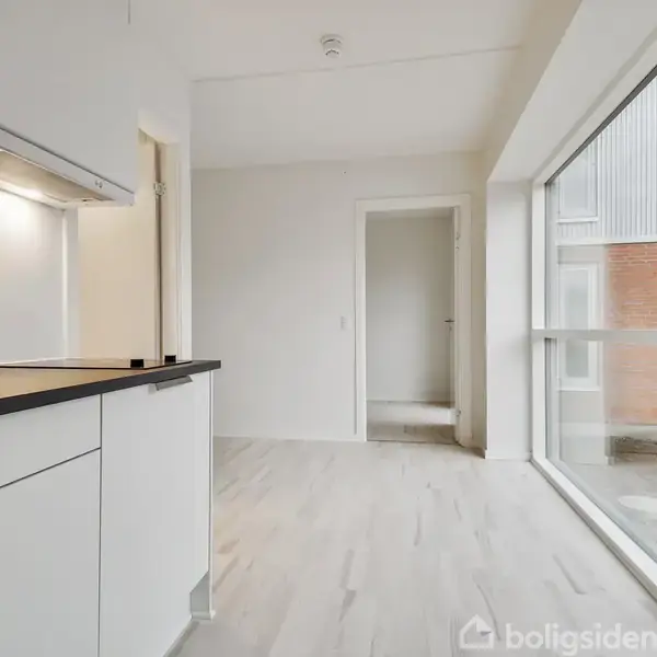 Et moderne køkken med hvide skabe og sort bordplade strækker sig langs en væg. Store vinduer til højre giver lys, og en dør åbner ind til et andet rum.