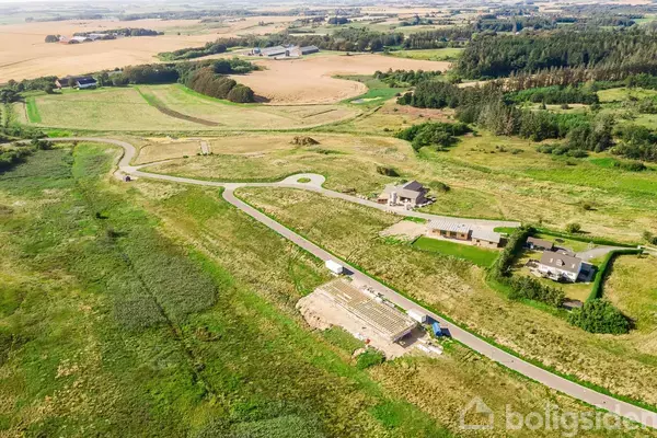 Luftfoto af et åbent landbrugsområde med spredte huse. Veje snor sig gennem grønne marker omgivet af træer. Distant bjerge kan skimtes i baggrunden under en lys himmel.
