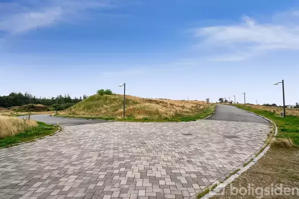 Brolagt vejstrækning fører op til skråning dækket af græs med lygtepæle langs vejen. Blanding af åben himmel og træbevoksede områder i baggrunden.
