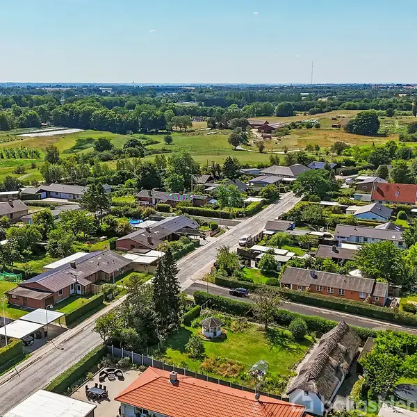 Boligområde med flere huse ligger langs en vej, som strækker sig gennem en grøn landskab af marker og træer. Solen skinner klart, og himlen er blå med få skyer.