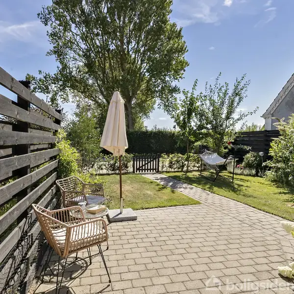 Terrasse med to stole og en parasol på stenbelægning, omgivet af frodig have med græs, blomster, et hegn og en hængekøje. En lille sti fører til en port.