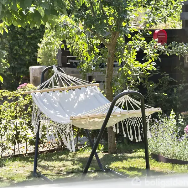 En hængekøje står stille i en solrig have, omgivet af grønne træer og blomsterbede. Det ser fredeligt og afslappende ud, med lysindfald, der skaber skygger på græsset.