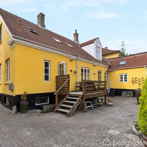 Et gulmalet hus med rødt tegltag har en træterrasse med trapper, i en gårdsplads af grå fliser. Plant dekoration står langs huset, mens en høj busk er synlig i højre side.