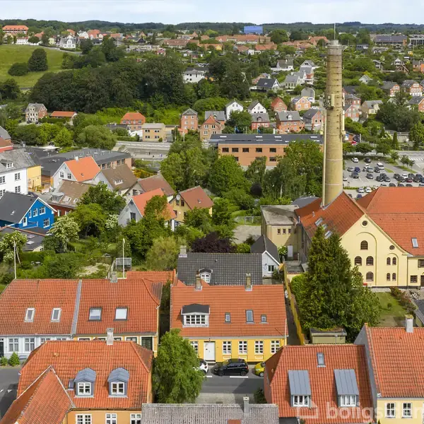 Bygninger med røde tage står stille i en by omgivet af grønne bakker og skove. En høj skorsten rager op midt i bebyggelsen, mens flere boliger spredes i det omkringliggende landskab.