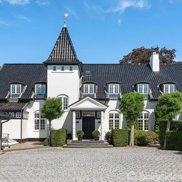 En stor, hvid villa med sort tag står i centrum. Den har en spiret tagkonstruktion. Foran er der en brolagt indkørsel og velplejede træer, omgivet af en åben port.