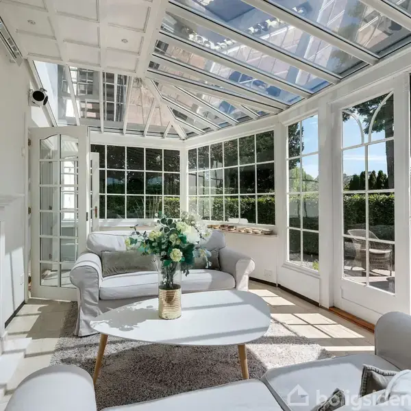En lys stue med glasvægge og loft, sofaer og et sofabord med blomster i en vase. Rummet åbner ud til en terrasse med haveudsigt gennem døre og vinduer.