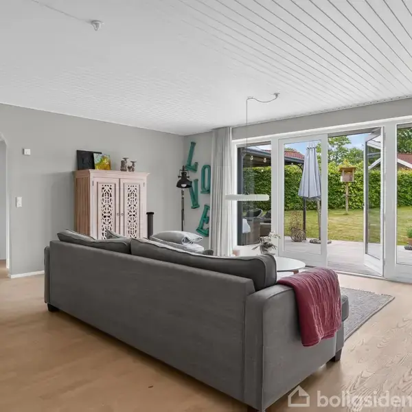 Grå sofa står i en stue med trægulv, vendt mod store glasdøre, der åbner ud til en terrasse og have. Et skab og dekorationen "LOVE" ses i hjørnet.
