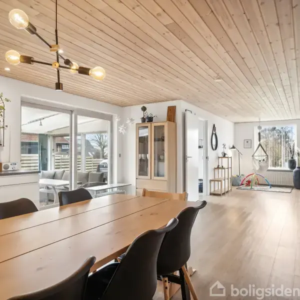 Langbord af træ med sorte stole, placeret i spisestue med lyst trægulv. Moderne loftlampe hænger over bordet. Stort vindue giver udsigt til udendørs terrasse. Hylder og skabe langs væggene.