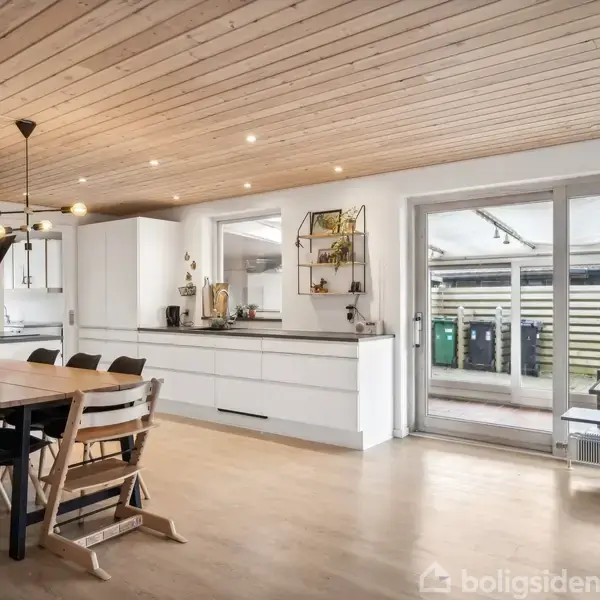 Et moderne køkken-alrum med spisebord og stole på trægulv. Køkkenskabe langs væggen, store vinduer til højre viser udendørs terrasse. Hængelamper giver belysning, væg har ur og kalender.