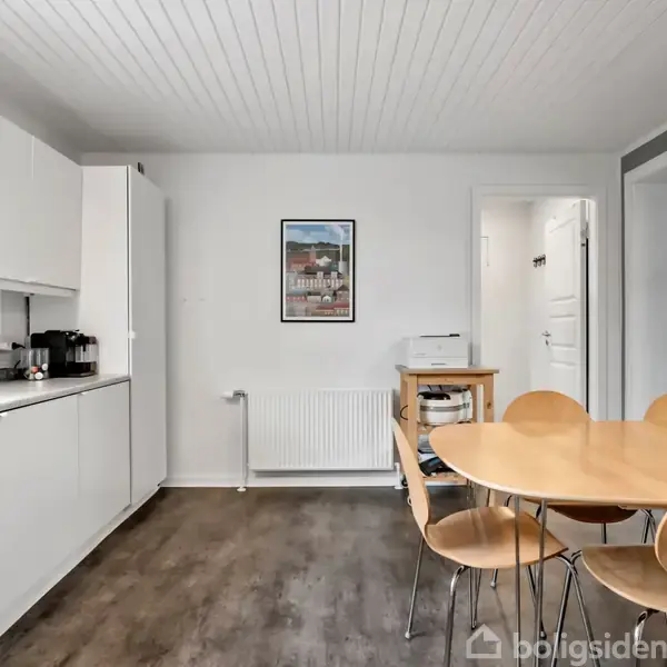 Et rundt træbord med fire stole står i et lyst køkken. Skabene er hvide, og gulvet er mørkt. En plakat hænger på væggen over en radiator.