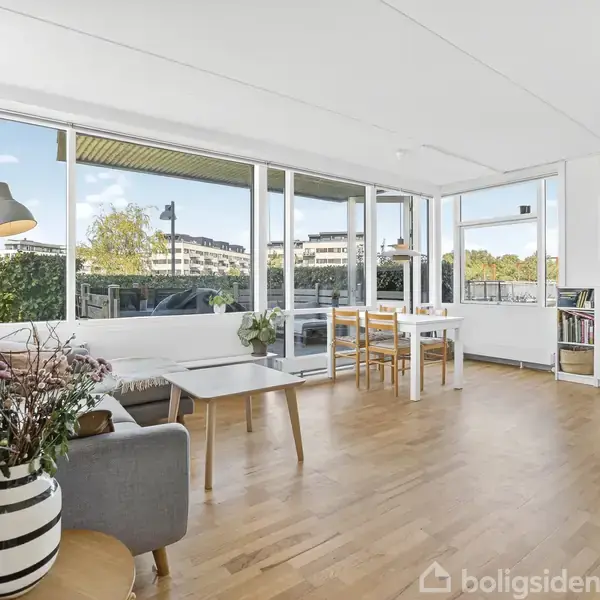 Stuen er lys med store vinduer, trælaminatgulv og moderne møbler. En grå sofa står til venstre, et hvidt bord i midten, og der er en bogreol til højre.