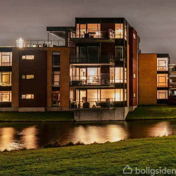 En lys stue med trægulv indeholder en grå sofa, et sofabord og en spiseplads ved vinduerne. Hylder med bøger og udsigt til grønne træer udenfor.