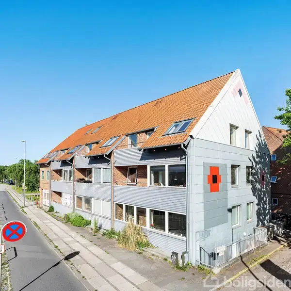 En bygning med rødt tegltag og flere vinduer langs facaden står ved en stille vej. Et vejskilt angiver parkeringsforbud. Der er træer og blå himmel i baggrunden.