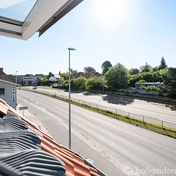Et åbent vindue på en bygning viser en fredelig, tom vej omkranset af grønne træer og huse i baggrunden. Solen skinner klart på en klar himmel.