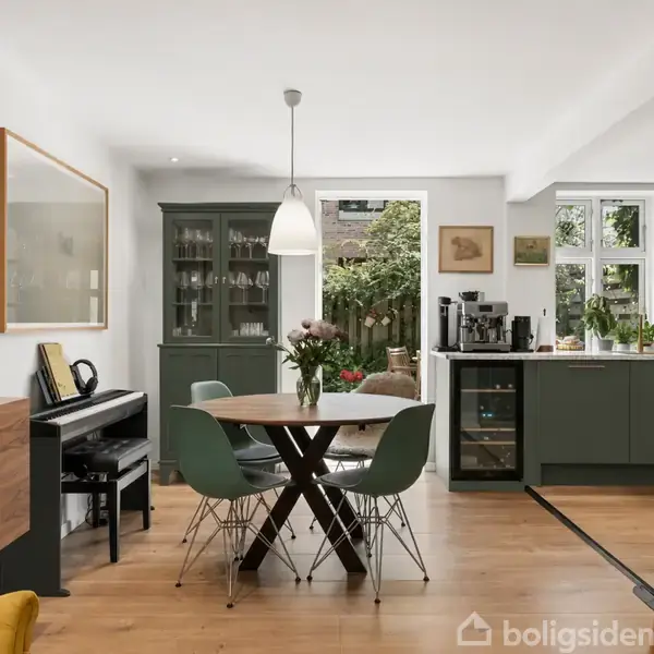 Et moderne køkken-alrum med et rundt spisebord midt i rummet, omgivet af grønne stole. Et klaver og malerier pryde væggene; store vinduer giver udsigt til en have.