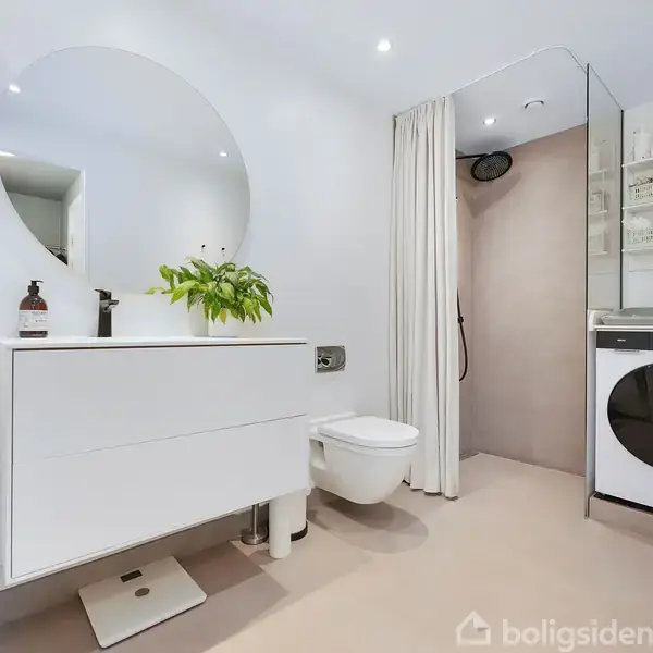 En moderne badeværelsesindretning med et rundt spejl over en hvid håndvask, hvorpå der står en plante og en sæbedispenser. Til højre ses et toilet og en vaskemaskine.