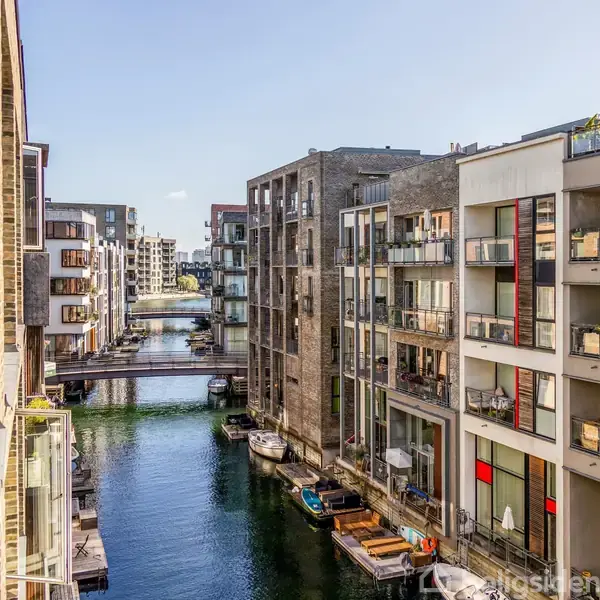 Bygninger flankerer en smal kanal, mens små både ligger fortøjret ved kajen. Moderne lejlighedskomplekser med altaner giver udsigt over vandet i en bymæssig kontekst.