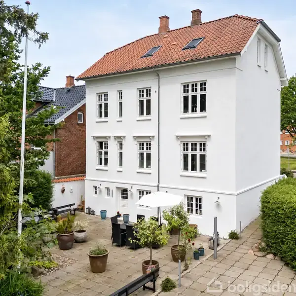 En hvid bygning med husnummeret 16 og en lanternelignende dekor står for enden af en trappe omkranset af hække. Til højre et glashus i en have omgivet af træer.