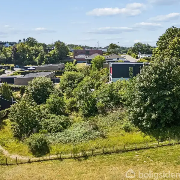 Boligområde med moderne huse omgivet af grønne træer og græs. Solrigt vejr og klar himmel skaber en fredelig atmosfære. Veje og stier forbinder de forskellige dele af landskabet.