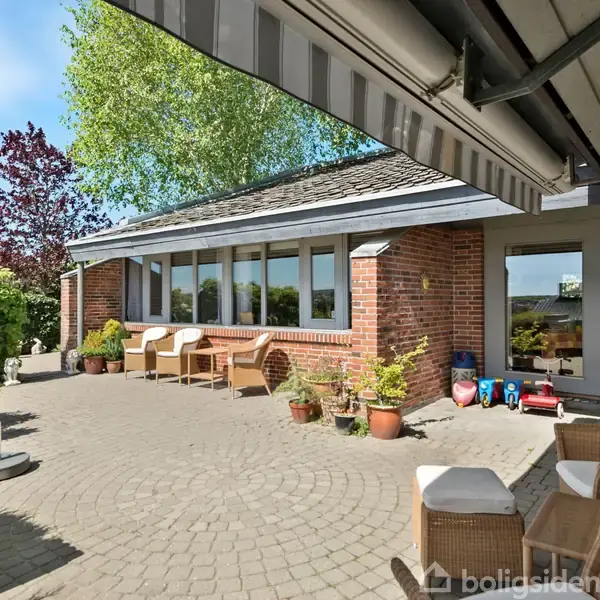 Terrasse med komfortable havemøbler hviler under en markise. Omkring står blomsterkrukker og legetøj, mens bagved ses et murstenshus med store vinduer, omgivet af grønne planter og klar himmel.