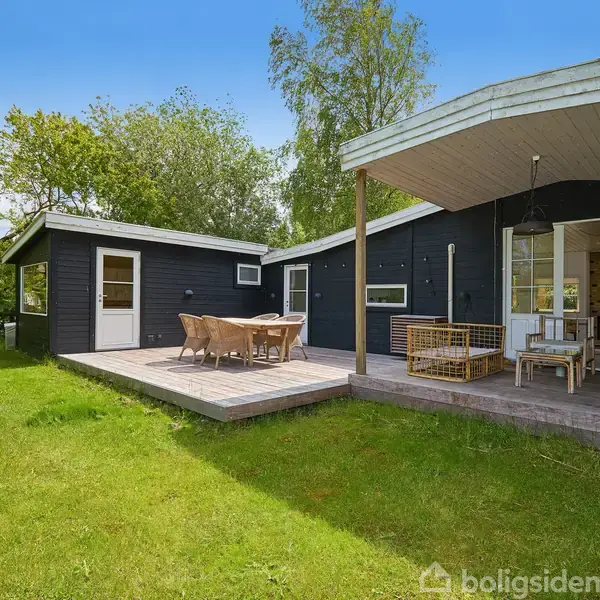 Sort træhus med åbne terrasser, hvor møbler står stillet op til afslapning. Huset er omgivet af grøn græsplæne og træer i en fredelig have.