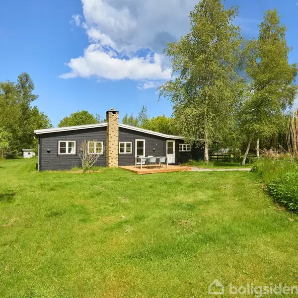 Sommerhus står stille omgivet af en grøn have med træer under en blå himmel med spredte skyer.