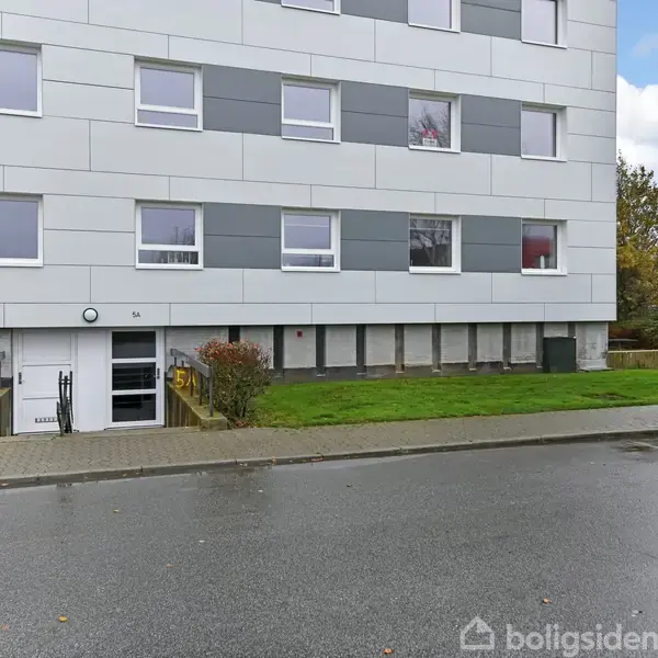 Et lejlighedskompleks med hvide og grå paneler har flere vinduer og en hovedindgang ved siden af en lille trappe. Foran er der græsplæne og fortov, omgivet af buske.