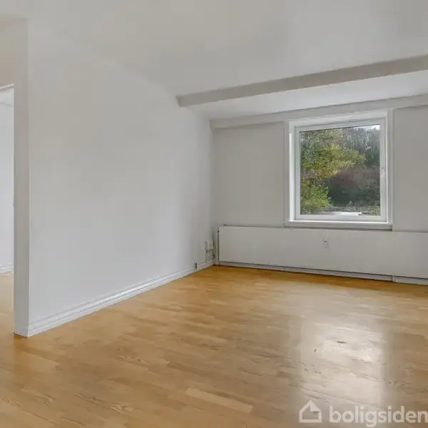 Et tomt rum med lyse trægulve og hvide vægge, vindue med udsigt til grønne træer, og en hvid radiator under vinduet; en åben dør fører til et tilstødende rum.