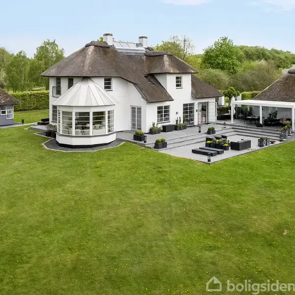 En stor, hvid villa med stråtag og udestue står på en velholdt græsplæne. Ved siden af ligger et mindre, lignende hus og en overdækket terrasse. Omkring ses grønne træer.
