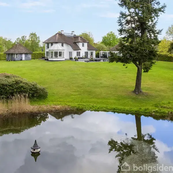 Stort hvidt hus med stråtag beliggende på en grøn, åben græsplæne. En lille dam i forgrunden reflekterer træer og himmel. Omkranset af buske og træer.