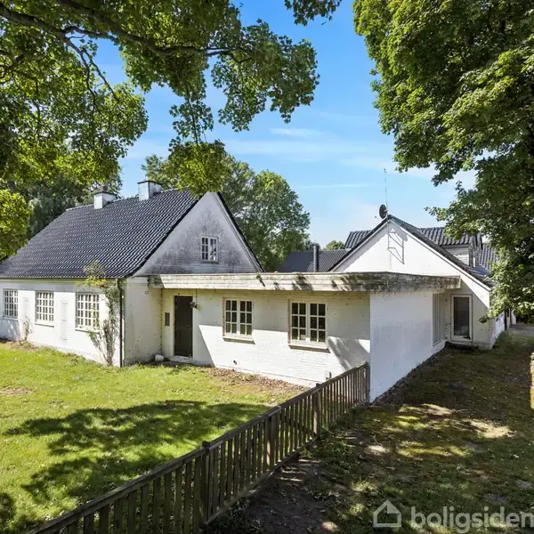 Hvidt hus med sort tag, omgivet af grøn beplantning og træer, står på en græsplæne med en lille træindhegning. Skyfri blå himmel i baggrunden.