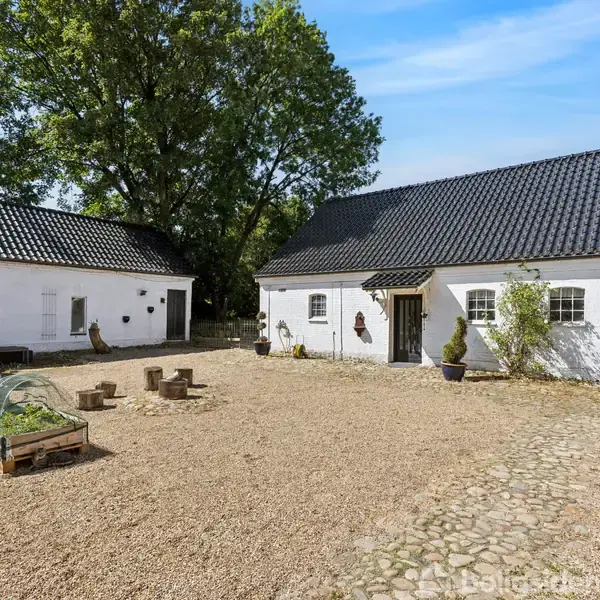 To hvidkalkede bygninger står omkring en grusgård med stengang. Der er plantekasser med grønne planter. Området er omgivet af træer under en blå himmel.