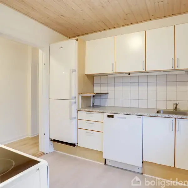 Et køkken med hvide skabe og en opvaskemaskine står sidstnævnte til venstre for vasken. Fliser på væggen og et køleskab ved siden af skabe. Lys trægulv og loft.