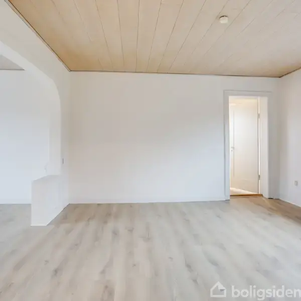 Et tomt rum med lyst trægulv og hvidmalede vægge. En indgang med buet døråbning fører til et tilstødende rum. Et vindue giver udsyn til det udendørs.