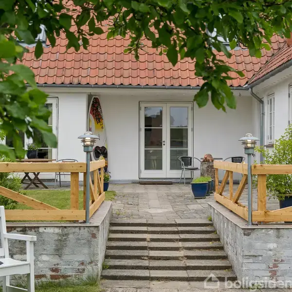 Hvid bænk står foran en hvid bygning med rødt tegltag. Terrasse med trægelænder er omgivet af grønne planter. Trappe fører op til glasdør, flankeret af lanterner.