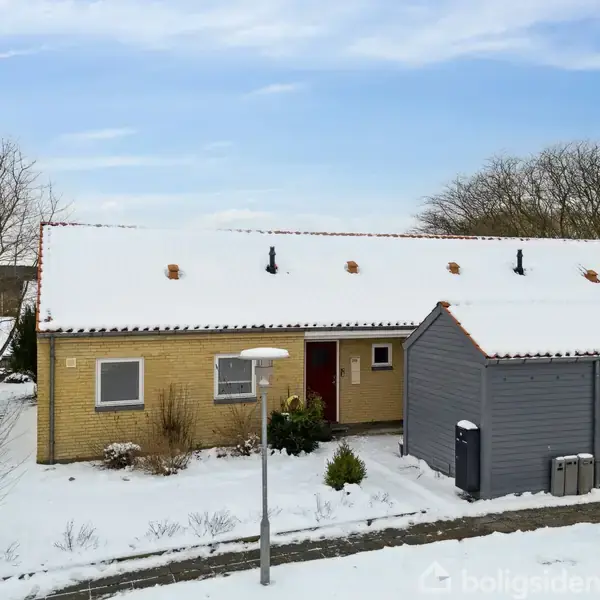 Hus dækket af sne står i en vinterlig have, omgivet af træer. Vejen foran er også snedækket. Der ses skorstene på det røde tegltag.