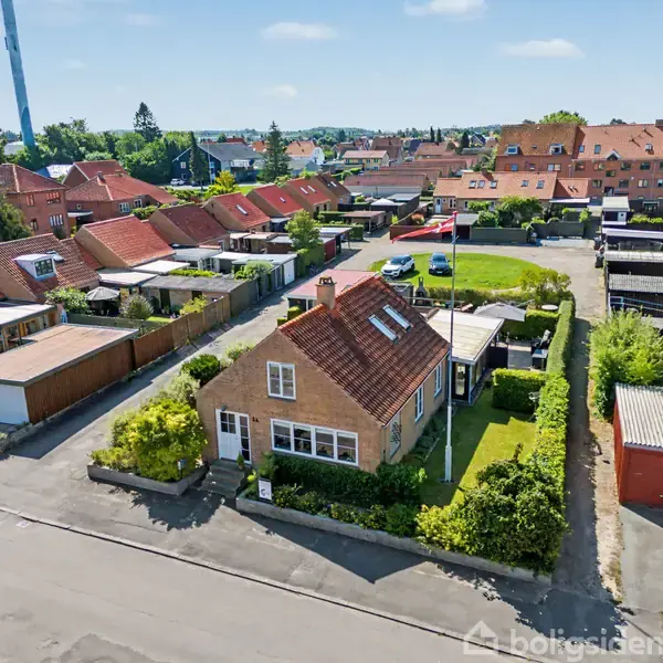 Et gult murstenshus med rødt tegltag står i et kvarter med lignende rækkehuse. Haven er velholdt med buske og græs. I baggrunden ses et parkområde og flere boliger.