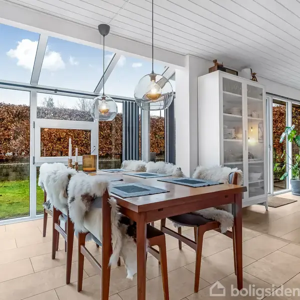 Spisebord dækket til fire med pelsbetrukne stole i en lys stue. Rummet har store vinduer mod en have med hæk. To moderne pendellamper hænger over bordet.