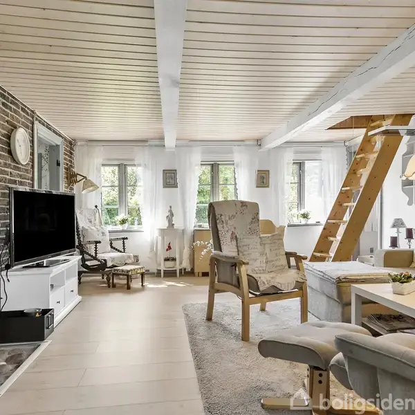 Stue med brændeovn op ad murstensvæg, der brænder ild; TV på skænk. Hyggelige møbler står på tæppe, trappe fører til hems. Vinduer giver udsigt til grønne omgivelser.