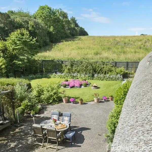 Et udendørs spisebord står på en terrasse omgivet af frodige buske og blomster. I baggrunden ses en grøn græsplæne og en lille bakke under en klar blå himmel.